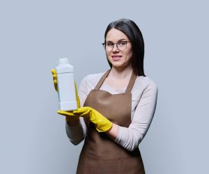 Young woman in gloves apron with organic detergent in hands looking at camera on light gray background. Home cleaning service, housekeeping cleanliness, natural eco organic detergents, ecology concept